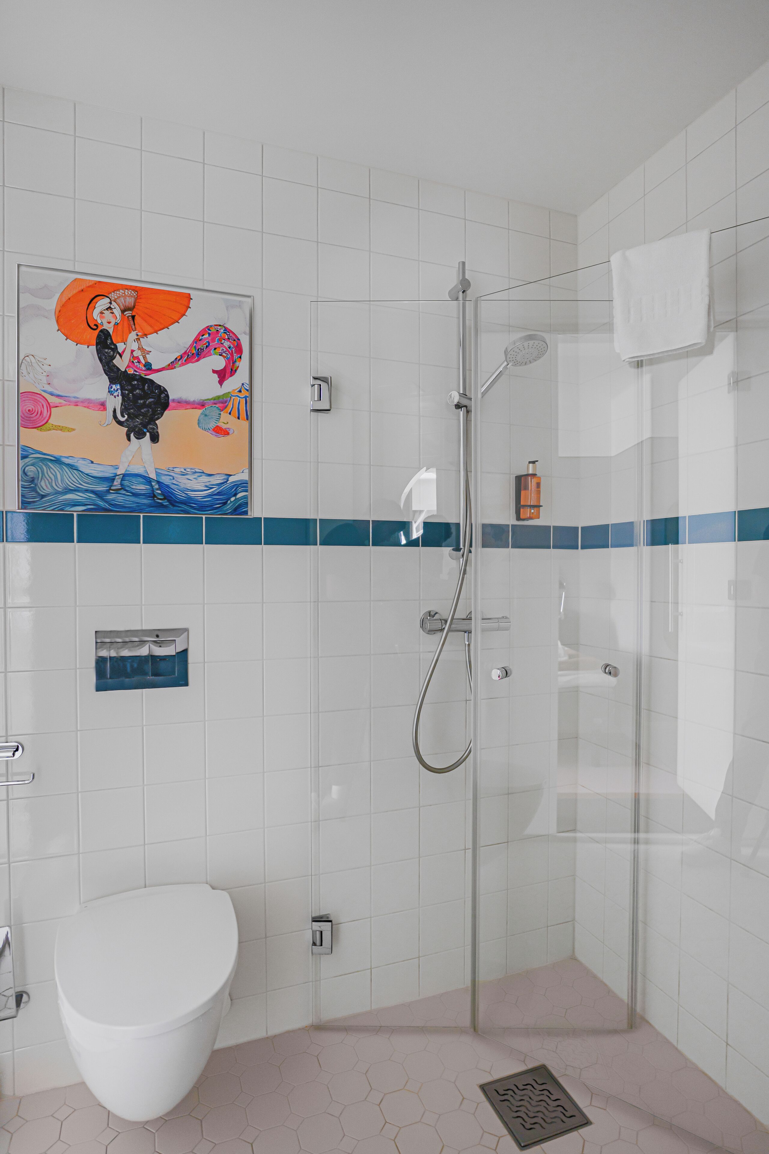A bathroom with a glass shower enclosure, a white toilet, and a tiled wall with a decorative painting.