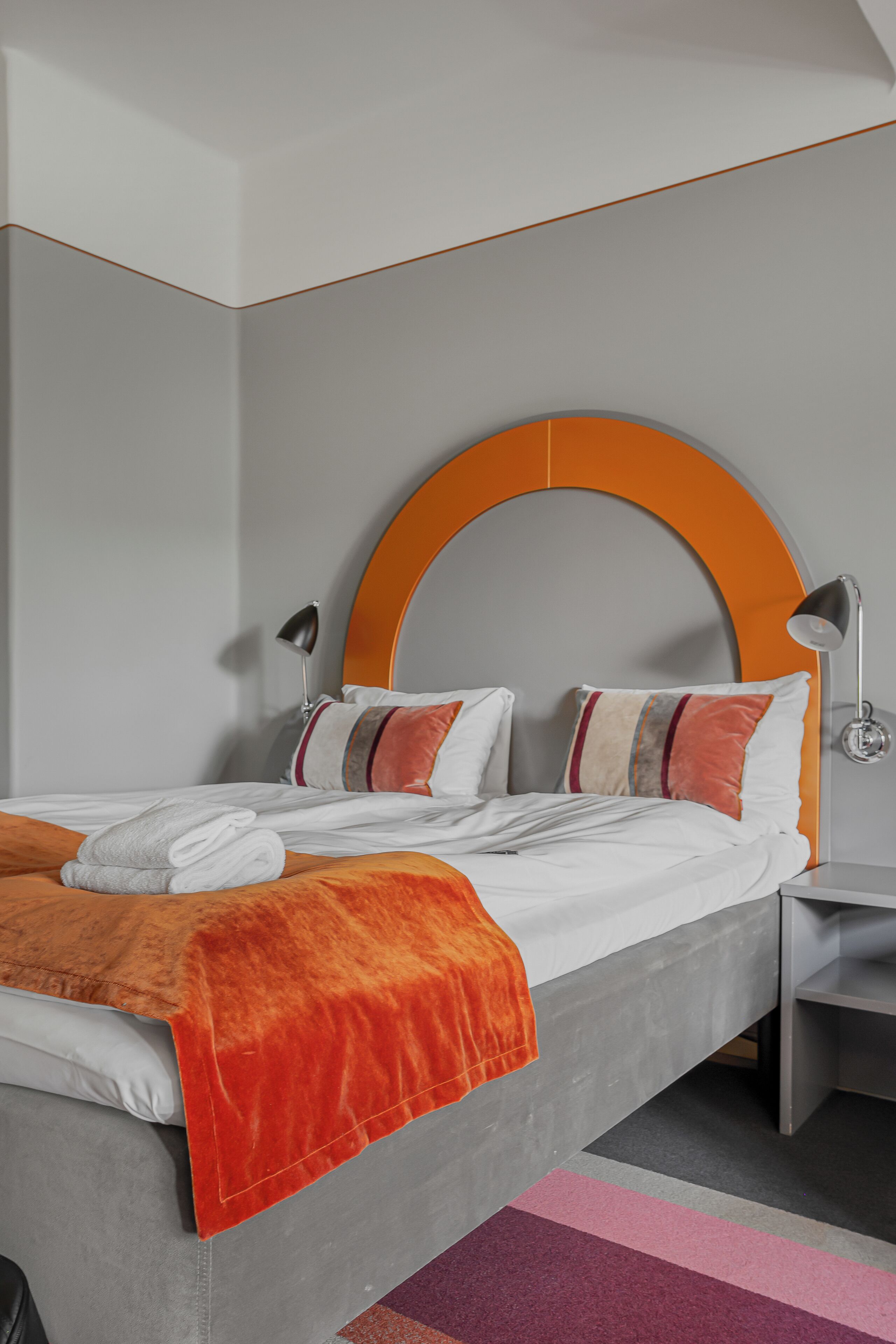 A modern hotel room with a grey bed, orange headboard, and a grey nightstand.