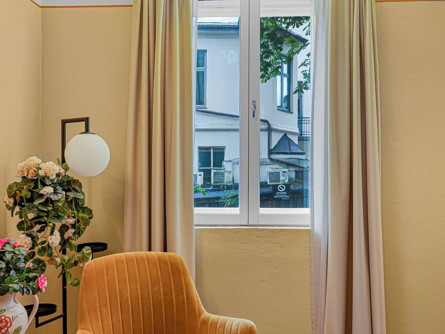 A room with a window, curtains, a chair, a lamp, and a floral arrangement.