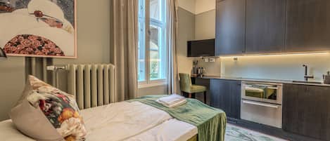 A bedroom with a bed, a green blanket, a pillow, a painting, a window with curtains, a radiator, a chair, and a kitchenette with a stove and sink.