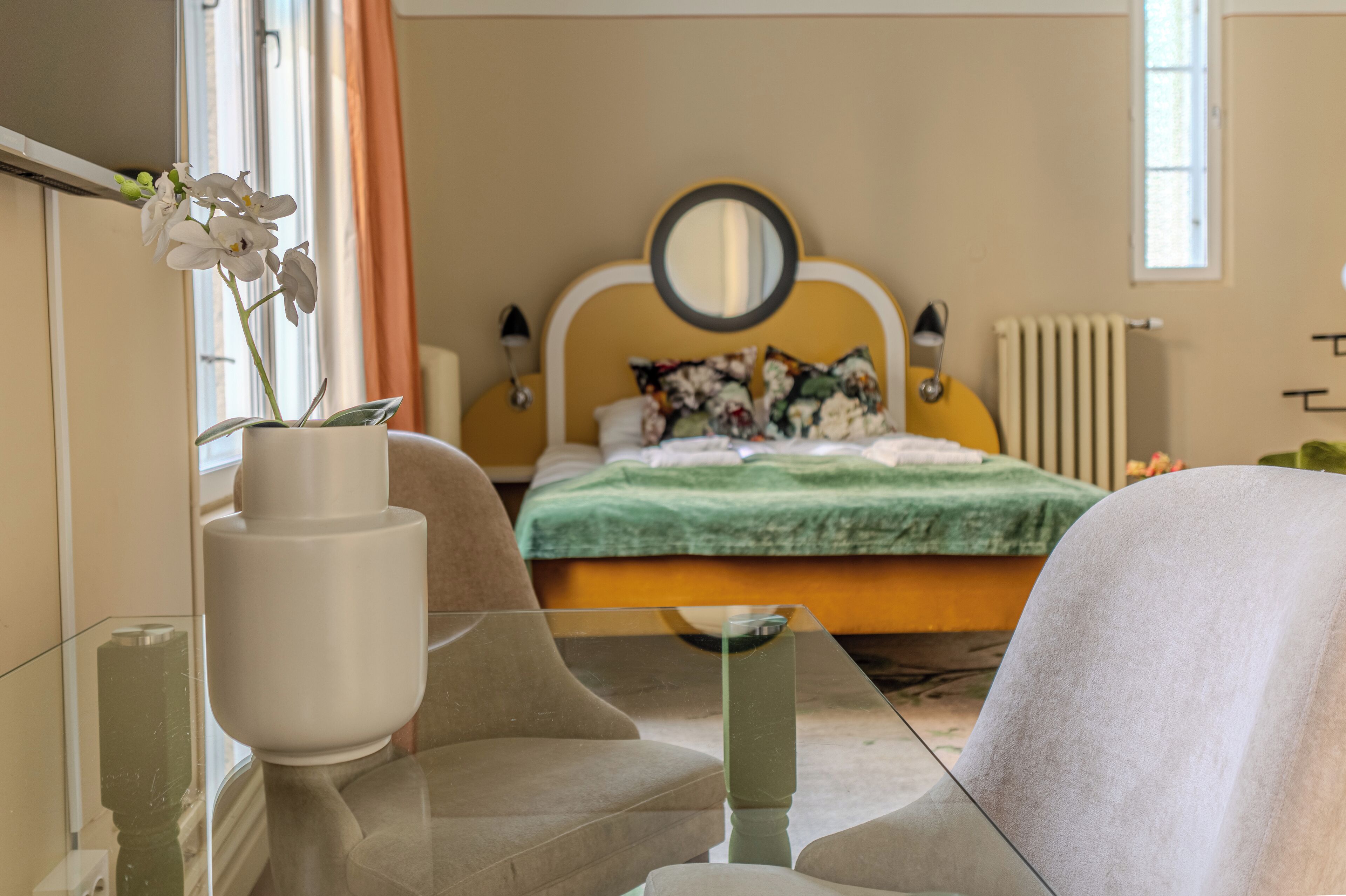 Ein Schlafzimmer mit einem Bett, einem Nachttisch, einem Sessel, einer Vase mit Blumen und einem Fenster.