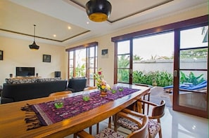 A dining area with a wooden table, chairs, and a purple table runner. There are large windows with a view of greenery outside.