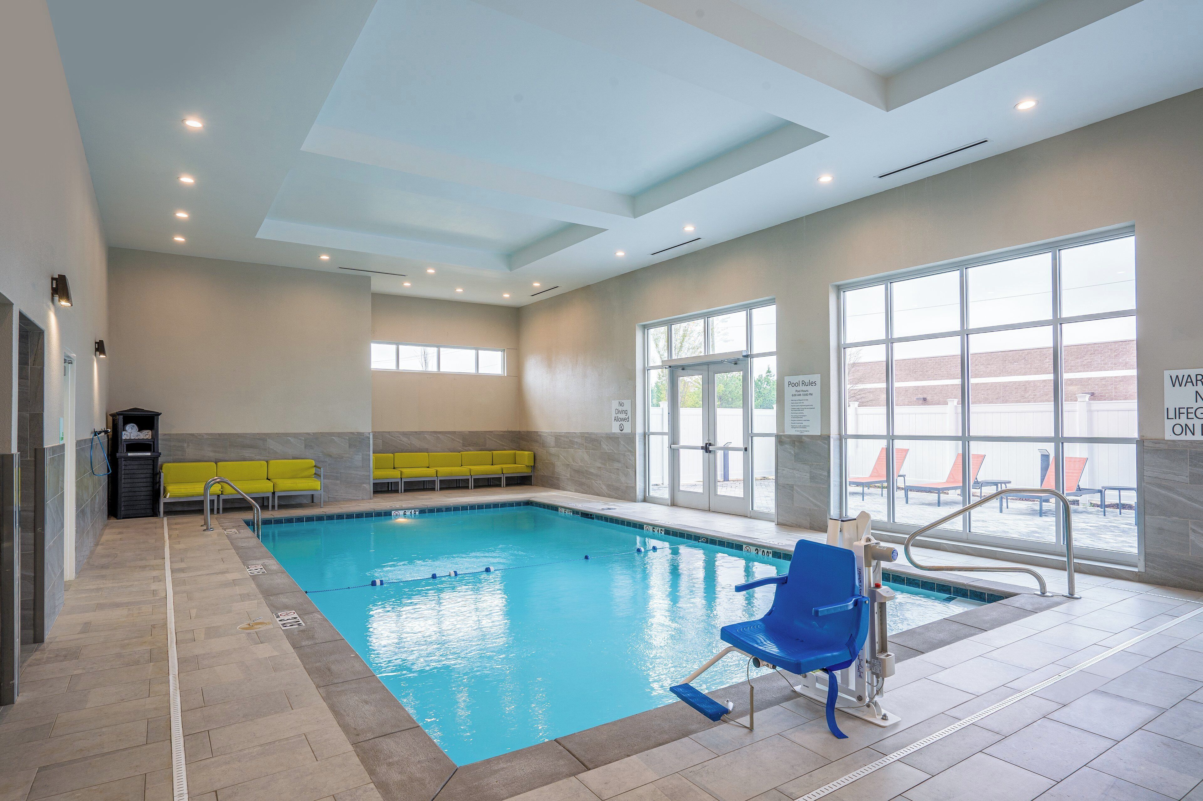 An indoor swimming pool with clear water, yellow and blue lounge chairs, and a poolside area with a view of the outside.