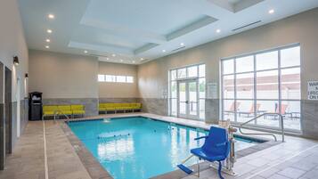 Una piscina interior con agua cristalina, sillas de descanso amarillas y azules, y un área junto a la piscina con vistas al exterior.
