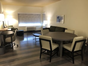 Un salon moderne avec une table à manger ronde, six chaises, un canapé, une table basse et une grande fenêtre avec des stores.