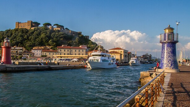 A coastal town with a prominent castle on a hill, a red lighthouse, and a white boat docked at the pier. - Yacht Suite Castiglione della Pescaia (Castiglione della Pescaia)
