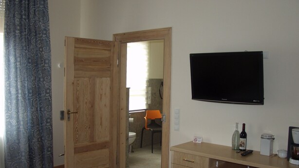 Ein Schlafzimmer mit Holztür, einem Bett, einem an der Wand befestigten Fernseher, einem Schreibtisch mit einer Weinflasche und einem Stuhl im Hintergrund.