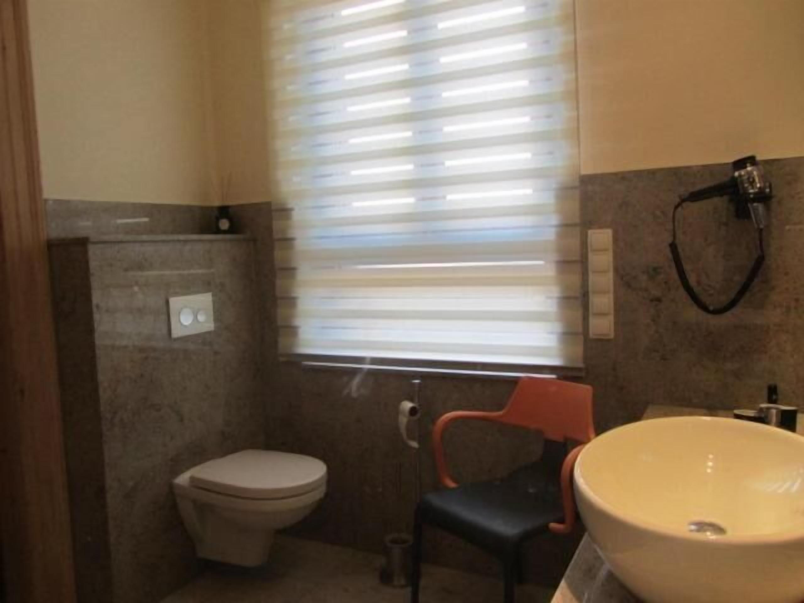 Ein Badezimmer mit Toilette, Waschbecken, einem Stuhl und einem Fenster mit Jalousien.