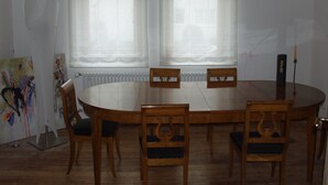Ein Esszimmer mit einem runden Tisch, sechs Holzsesseln, zwei Fenstern mit durchsichtigen Vorhängen und einem Heizkörper unter den Fenstern.