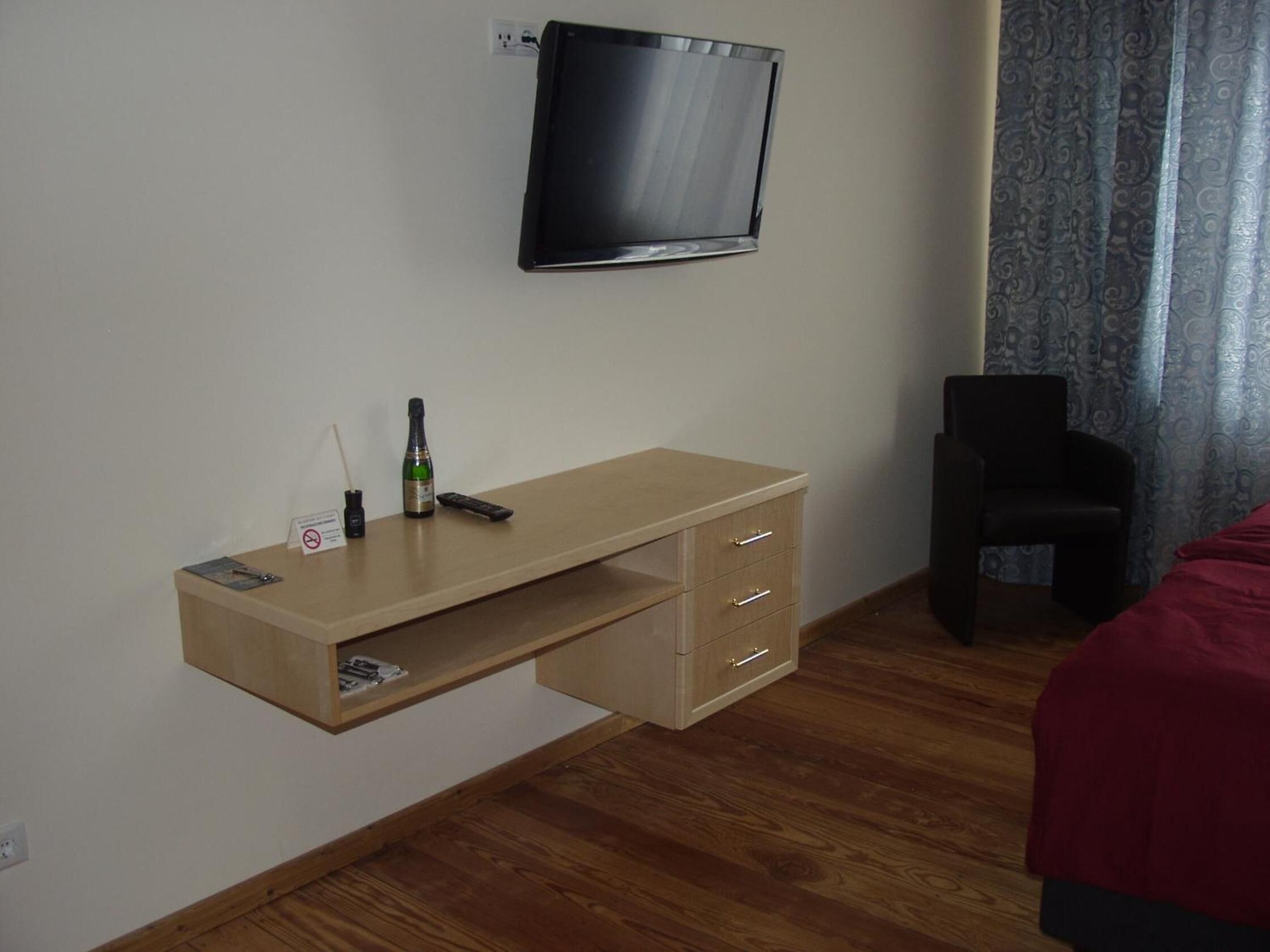 Habitación de hotel con un escritorio de madera, un televisor montado en la pared, una silla y una cama con cubrecolcha rojo.