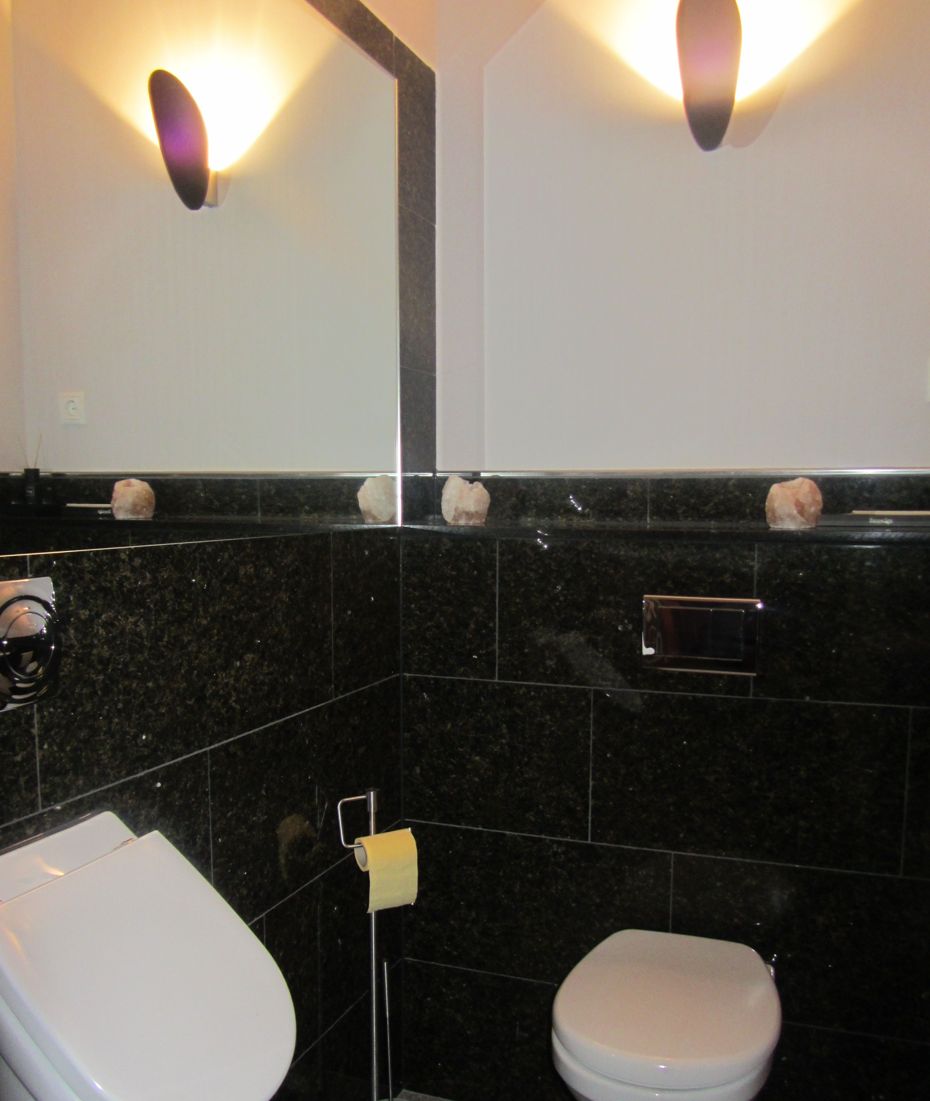 A bathroom with a white toilet, a sink, and a mirror. There are two wall-mounted lights and a towel dispenser.