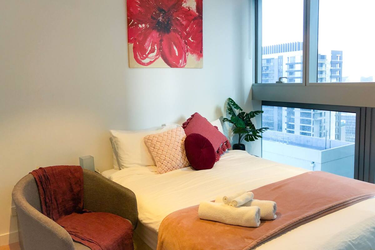 A hotel room with a bed, a large window, a red flower painting, and a plant.