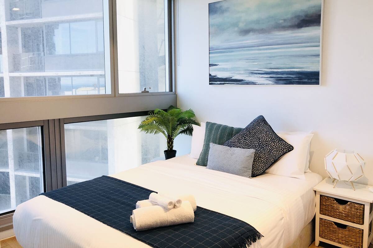 A modern bedroom with a large window, a bed with a dark quilted bedspread, a potted plant, and a wall-mounted abstract painting.