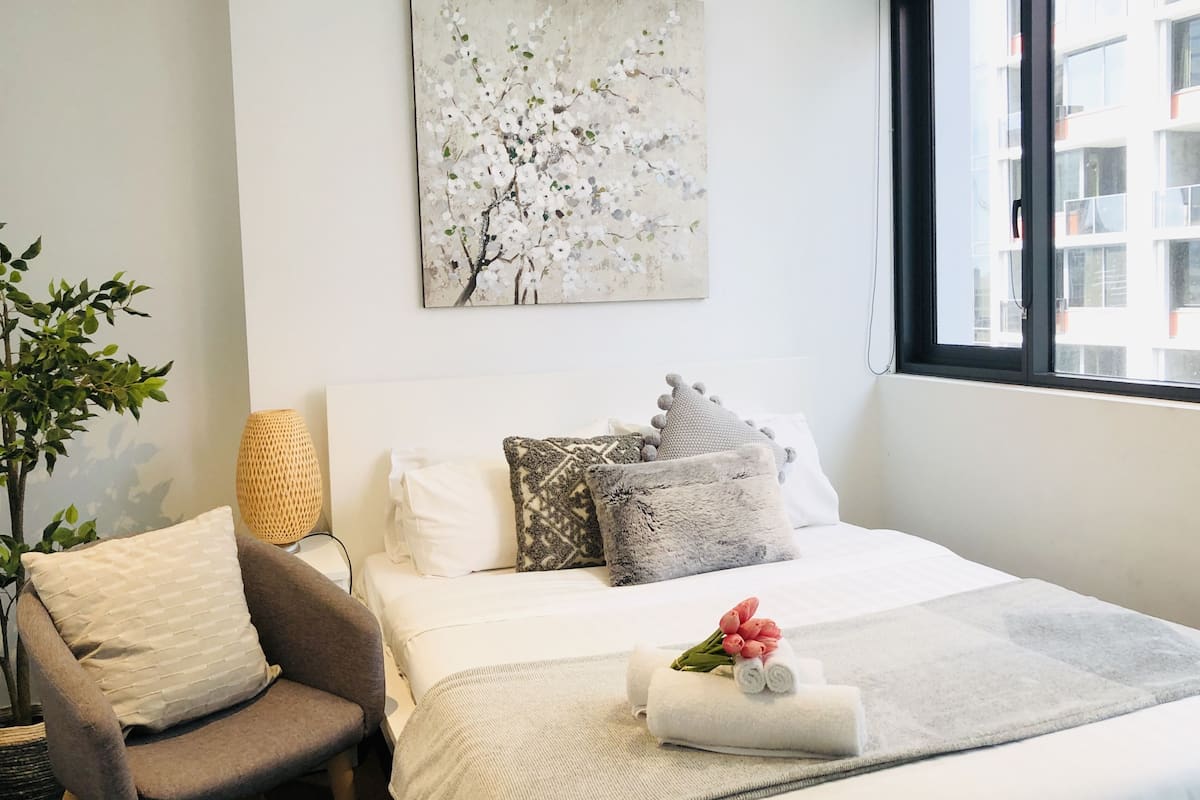 A hotel room with a bed, a chair, a potted plant, and a window with a view of buildings.