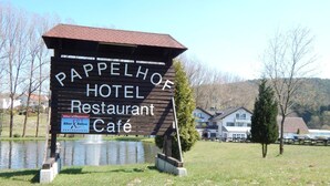 A sign for Pappelhof Hotel Restaurant Café with a wooden structure and a pond in the background. - Hotel Pappelhof (Weidenbach)