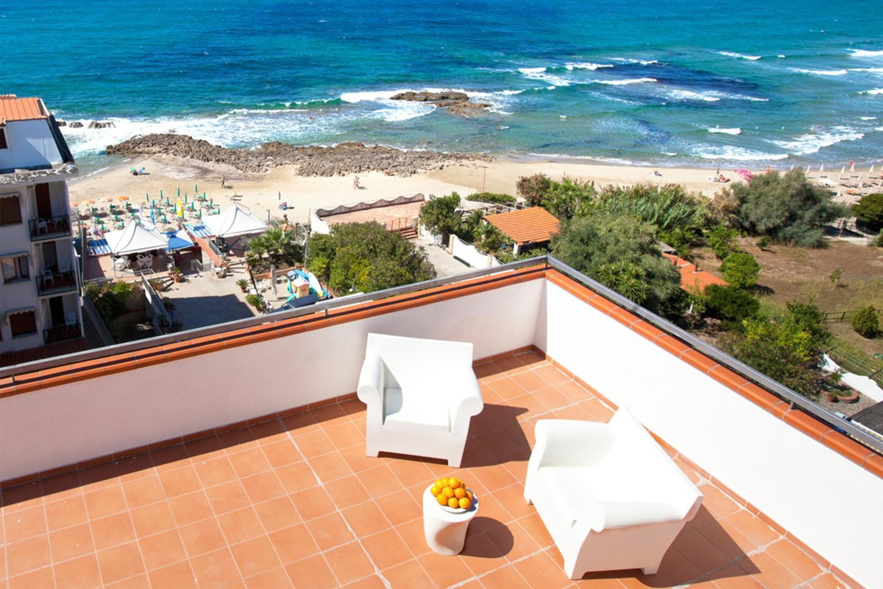 Un balcon avec des chaises blanches et une petite table ornée d’oranges, donnant sur une plage parsemée de parasols et animée par la présence de nombreuses personnes.