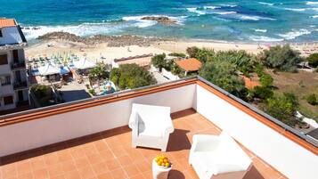 Un balcon avec des chaises blanches et une petite table ornée d’oranges, donnant sur une plage parsemée de parasols et animée par la présence de nombreuses personnes.