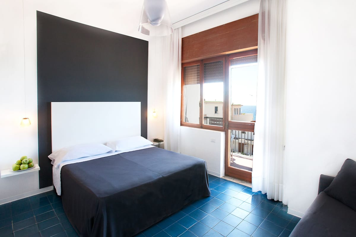 A hotel room with a bed, a chair, a nightstand, and a window with a view of the sea.