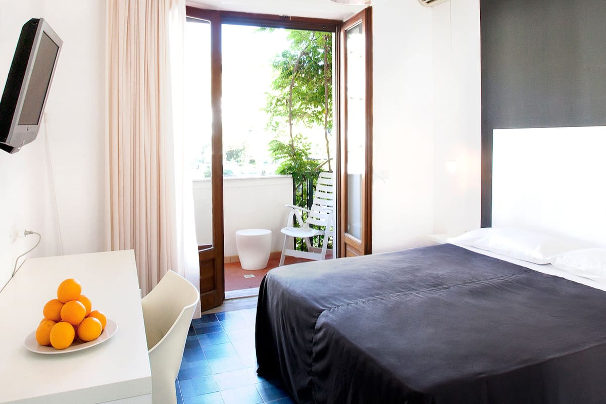A hotel room with a bed, a desk with a plate of oranges, a chair, and a view of the outdoors.