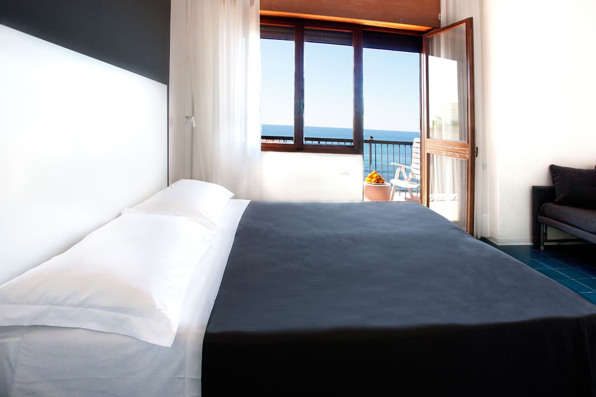 A hotel room with a bed, a sofa, a balcony with a view of the sea, and a potted plant.