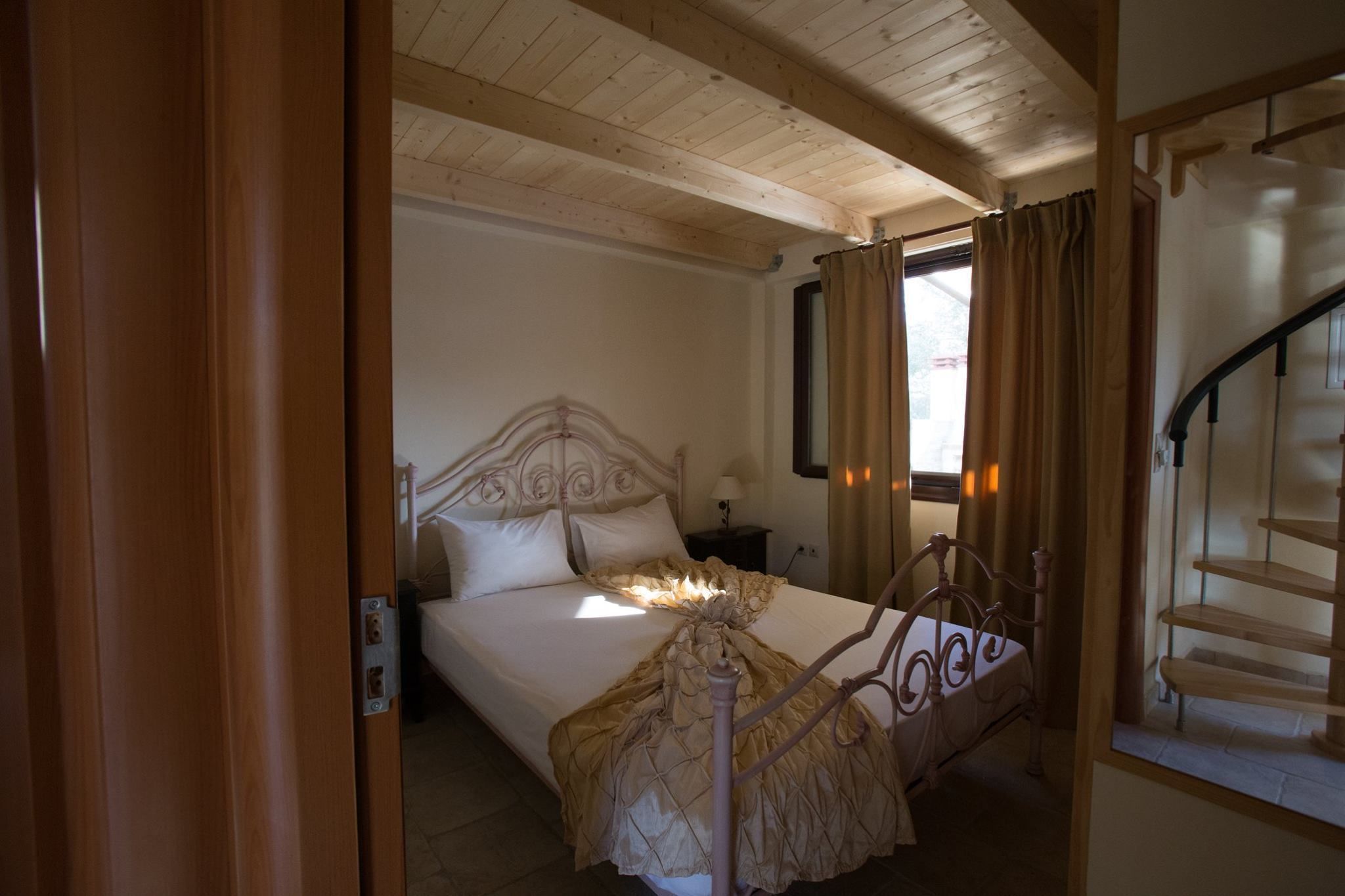 A bedroom with a wooden ceiling, a bed with a decorative headboard, a nightstand, and a staircase to the right.