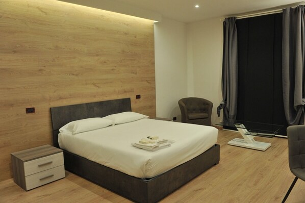 Una habitación de hotel moderna con un cabecero de madera, una cama individual con ropa blanca, una mesita de noche, una silla y una ventana con cortinas.