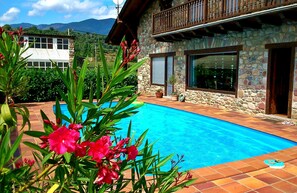 Um edifício de pedra com piscina, cercado por vegetação e montanhas ao fundo.