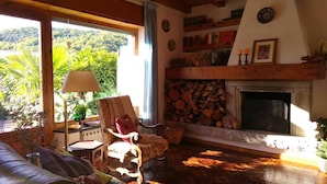 Un salon chaleureux avec une cheminée, un fauteuil confortable, un canapé et une vue sur une végétation luxuriante.