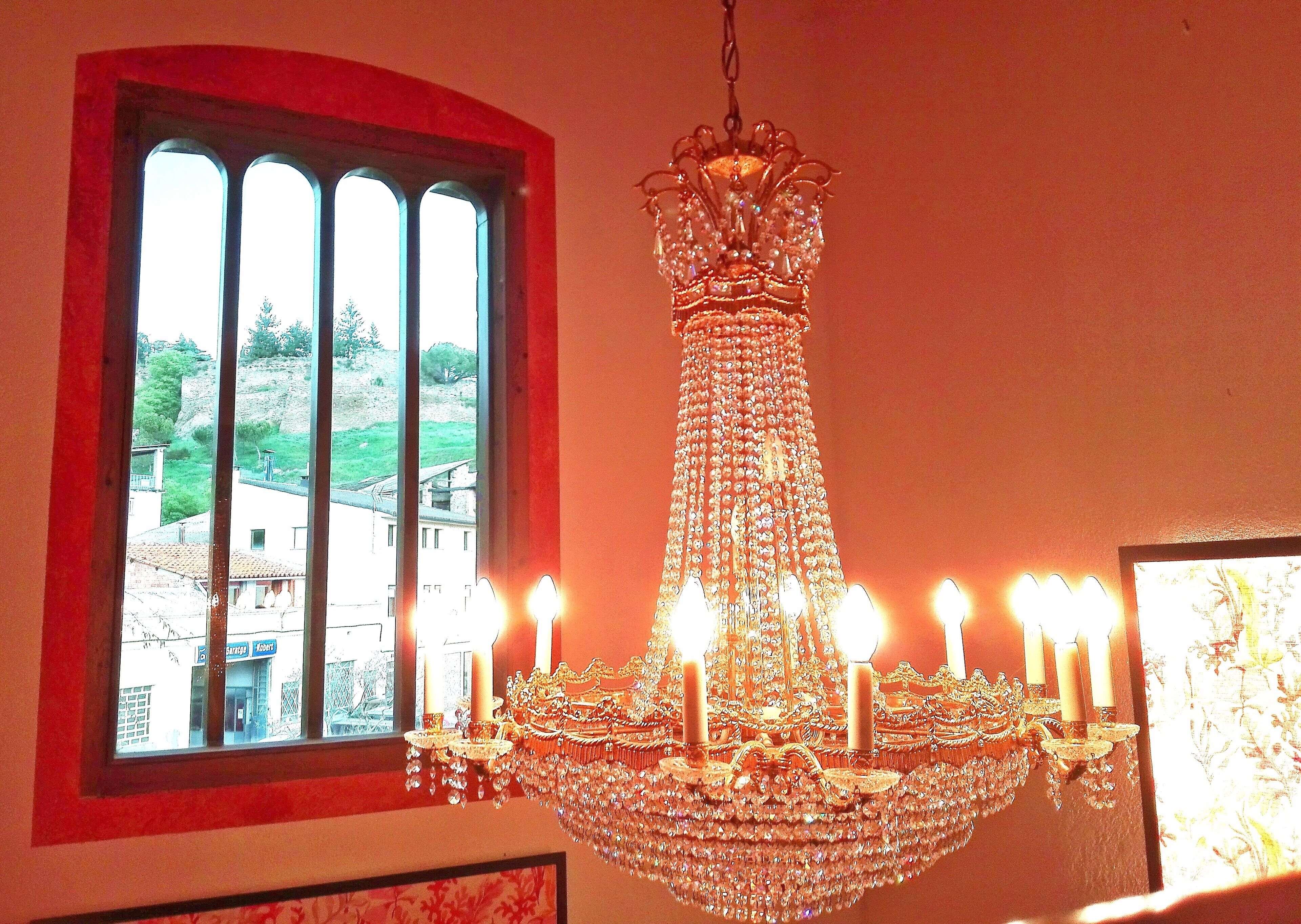 A chandelier with multiple candles, a window with a view of buildings, and a framed picture on the wall.