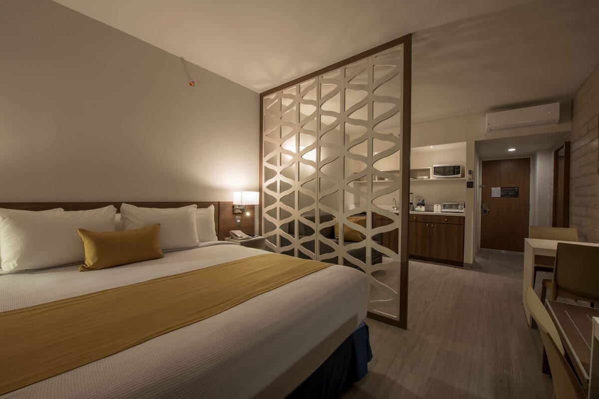 A hotel room with a large bed, a wooden headboard, a decorative partition, a bedside table with a lamp, and a kitchenette in the background.