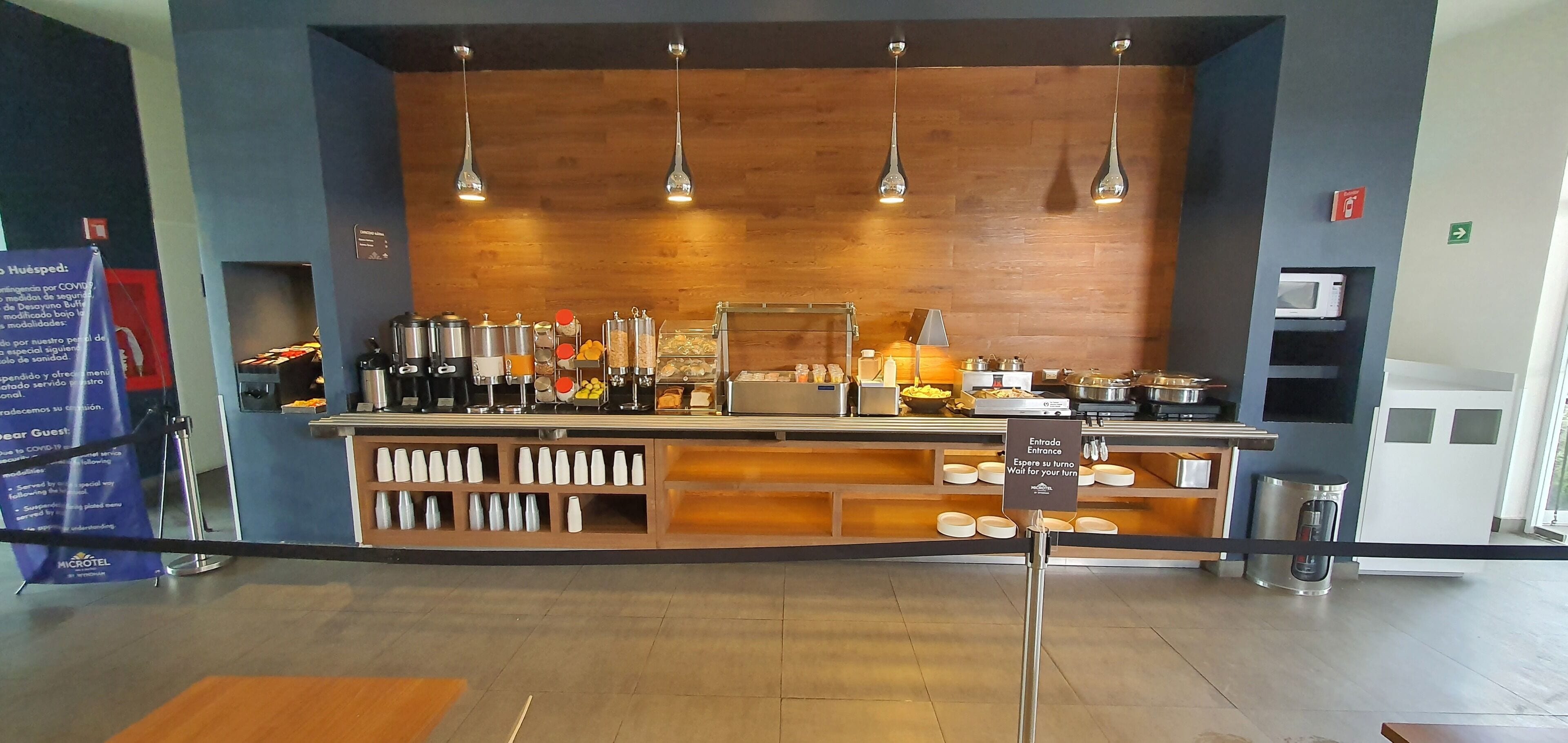 A hotel reception area with a counter, various food and beverage dispensers, a sign indicating the entrance, and a microwave.
