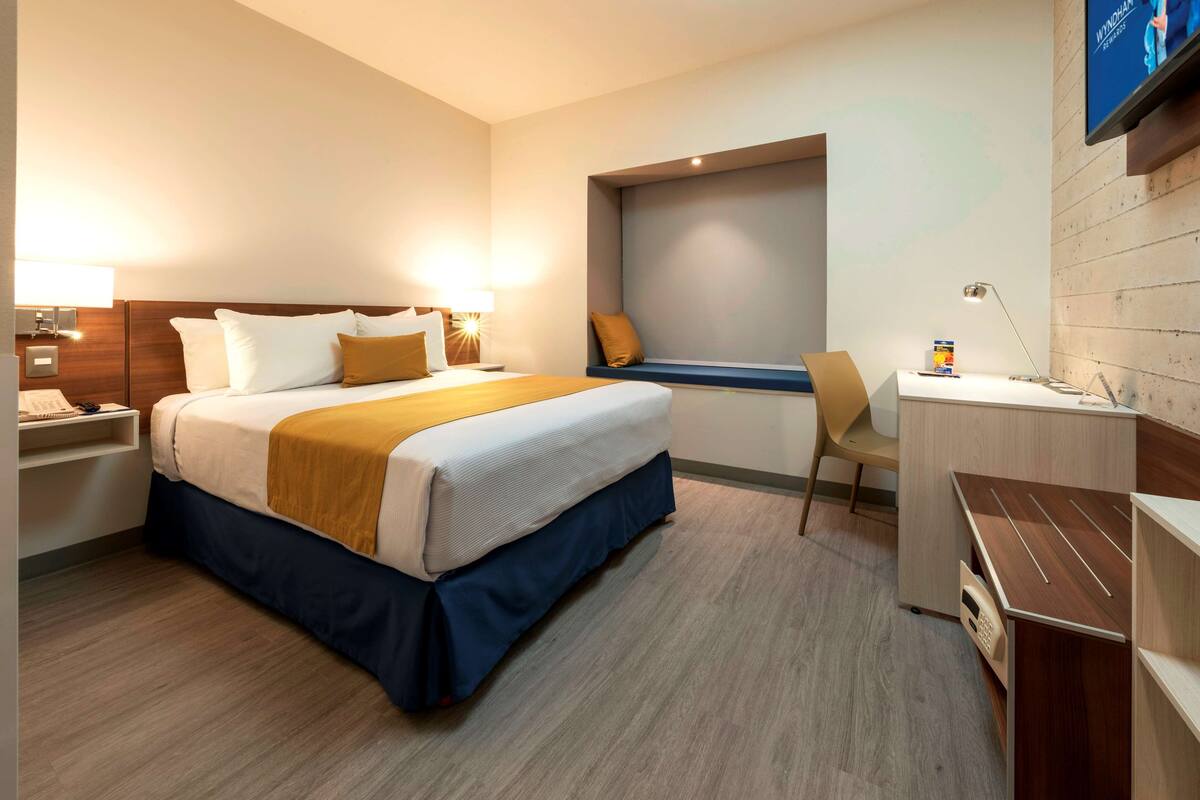 A hotel room with a bed, bedside table, desk, chair, and a TV mounted on the wall.