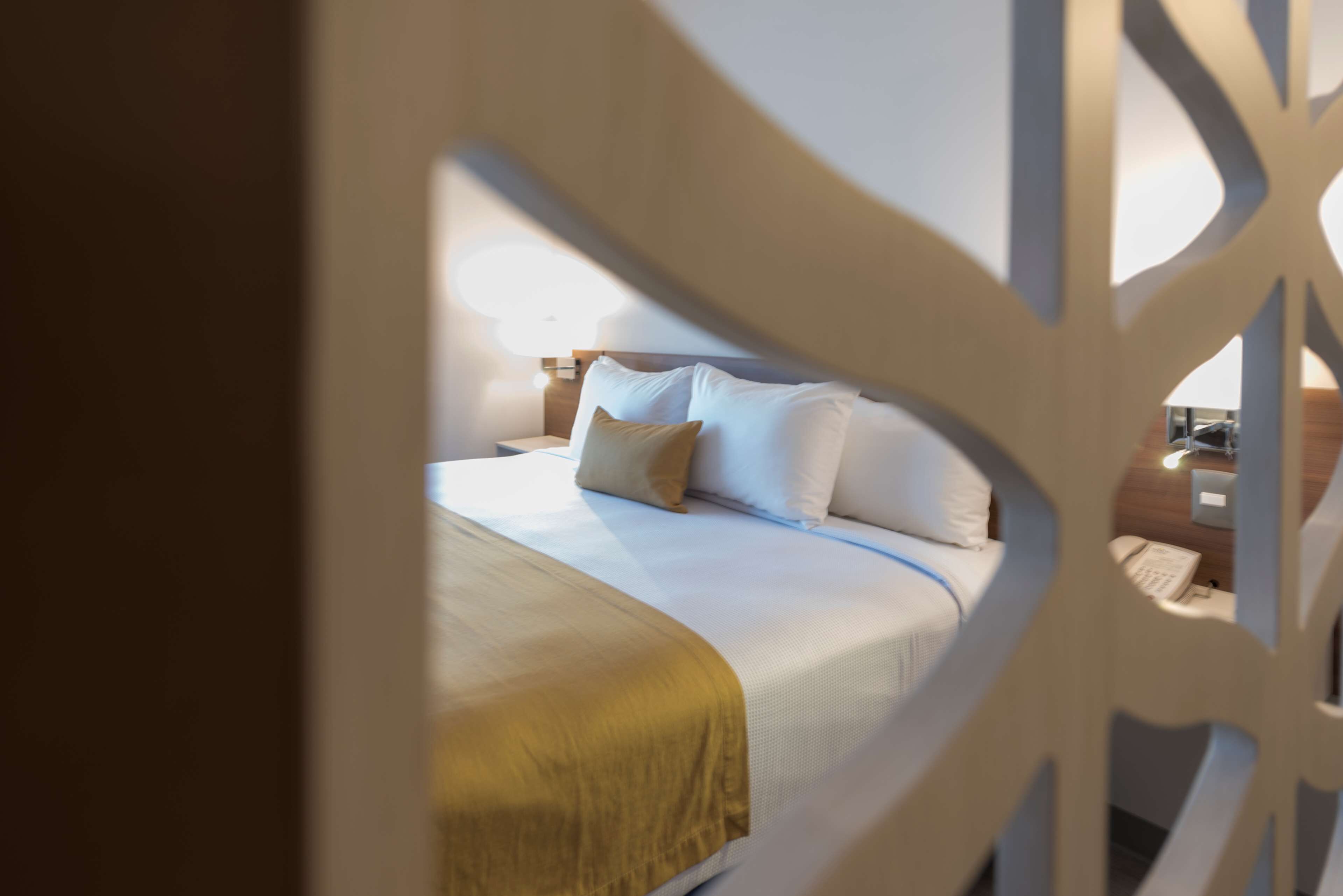 Una cama bien tendida con sábanas blancas y una manta dorada, una mesita de noche con lámpara y un cabecero de madera.