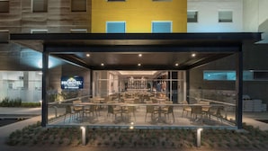 Exterior de un hotel moderno con fachada de vidrio, zona de estar al aire libre y el interior de un restaurante visible.