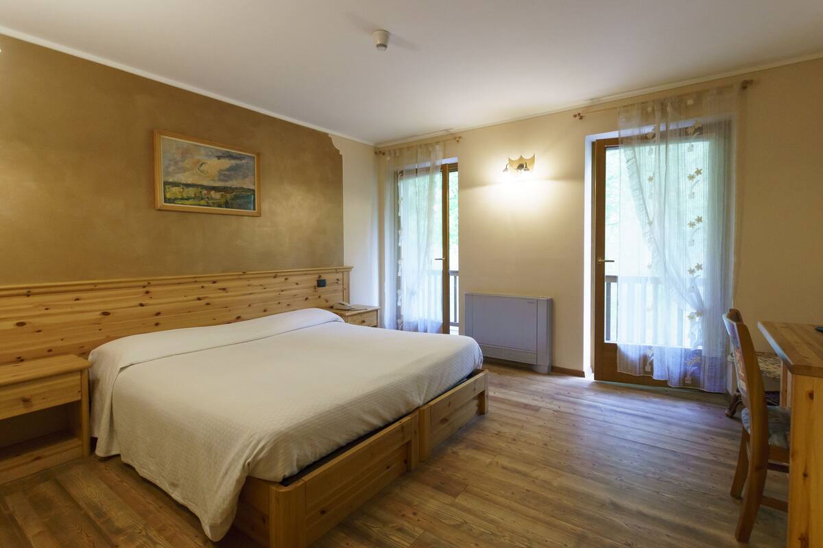 Un dormitori amb un llit de fusta, una tauleta de nit, un quadre a la paret i una finestra amb cortines translúcides.