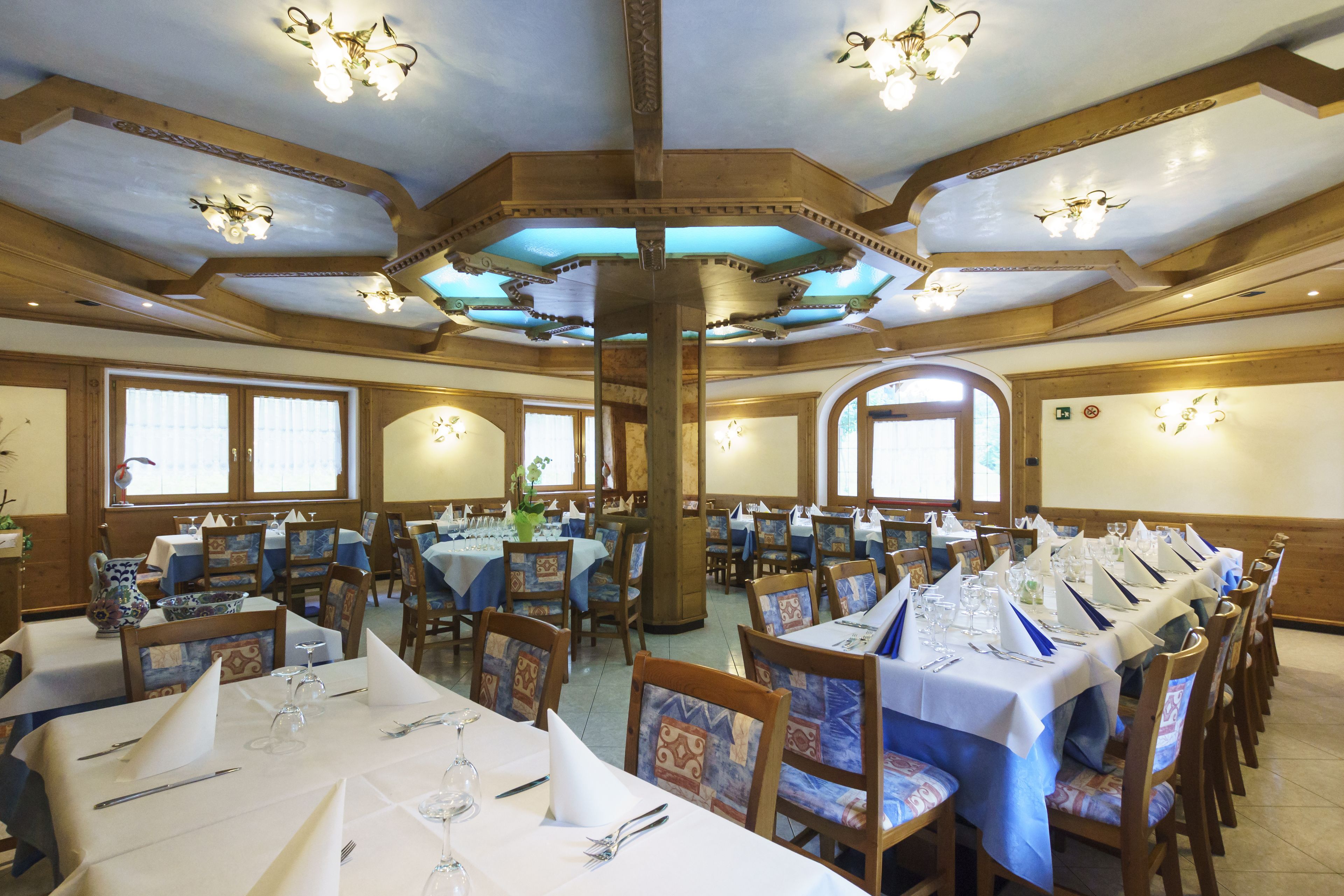 A dining hall with multiple tables set for a meal, wooden beams on the ceiling, and large windows allowing natural light.