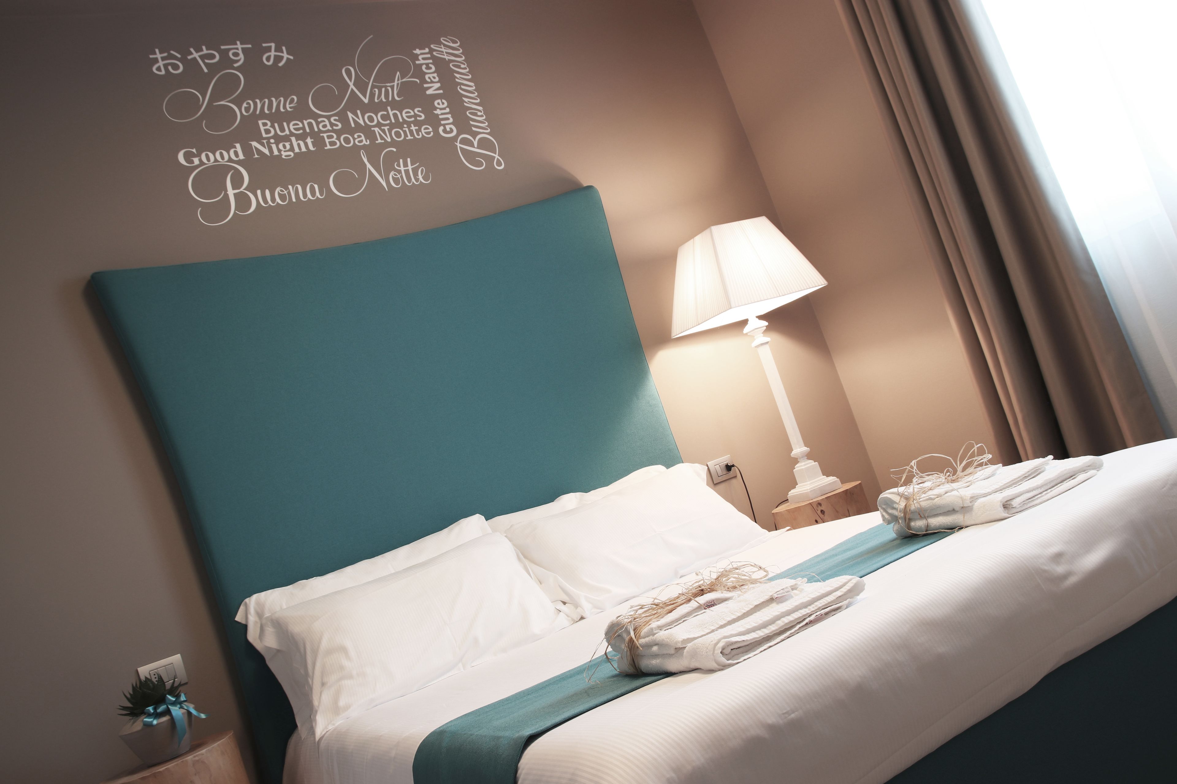 A neatly made bed with white linens and a teal headboard, a bedside lamp, and a wall decoration with multilingual good night wishes.