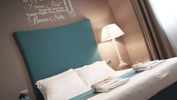 A neatly made bed with white linens and a teal headboard, a bedside lamp, and a wall decoration with multilingual good night wishes.