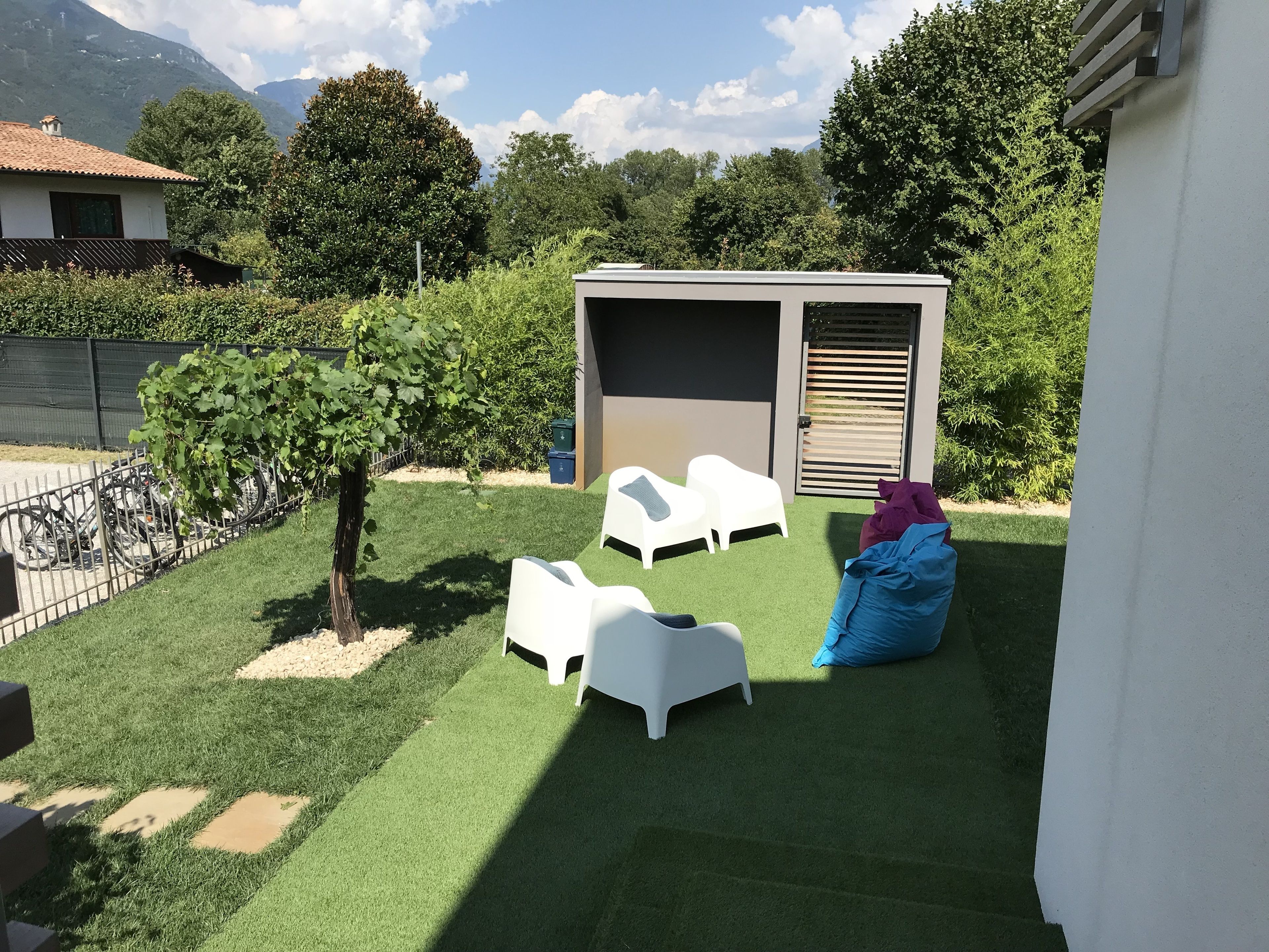 Un petit espace extérieur avec du gazon synthétique, des chaises en plastique blanches, un pouf bleu et un abri de jardin.