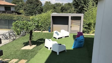 Un petit espace extérieur avec du gazon synthétique, des chaises en plastique blanches, un pouf bleu et un abri de jardin.