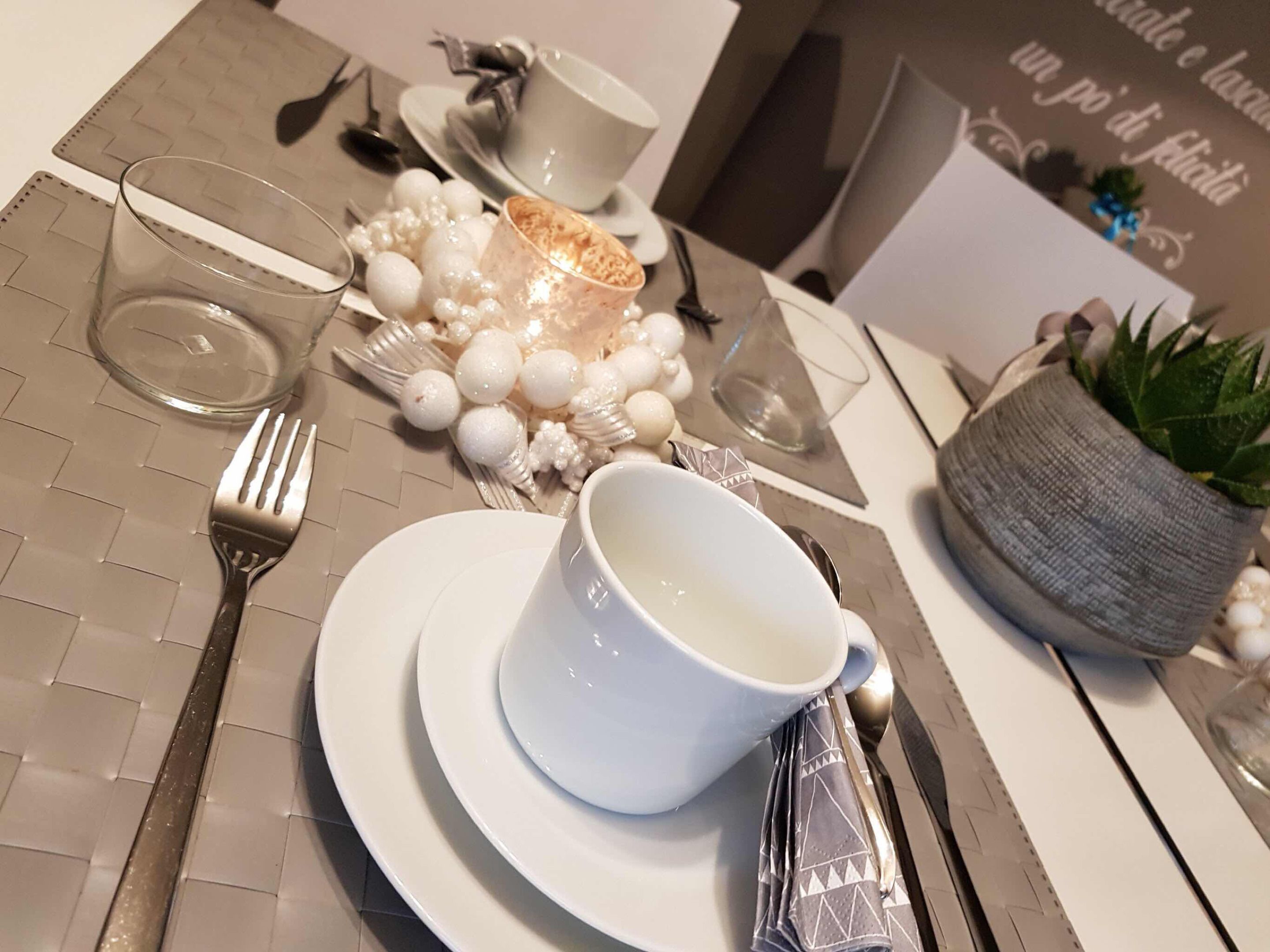 A neatly arranged dining table with a white cup, saucer, and glass, a fork, a knife, and a napkin. There is a small potted plant and decorative white spheres on the table.