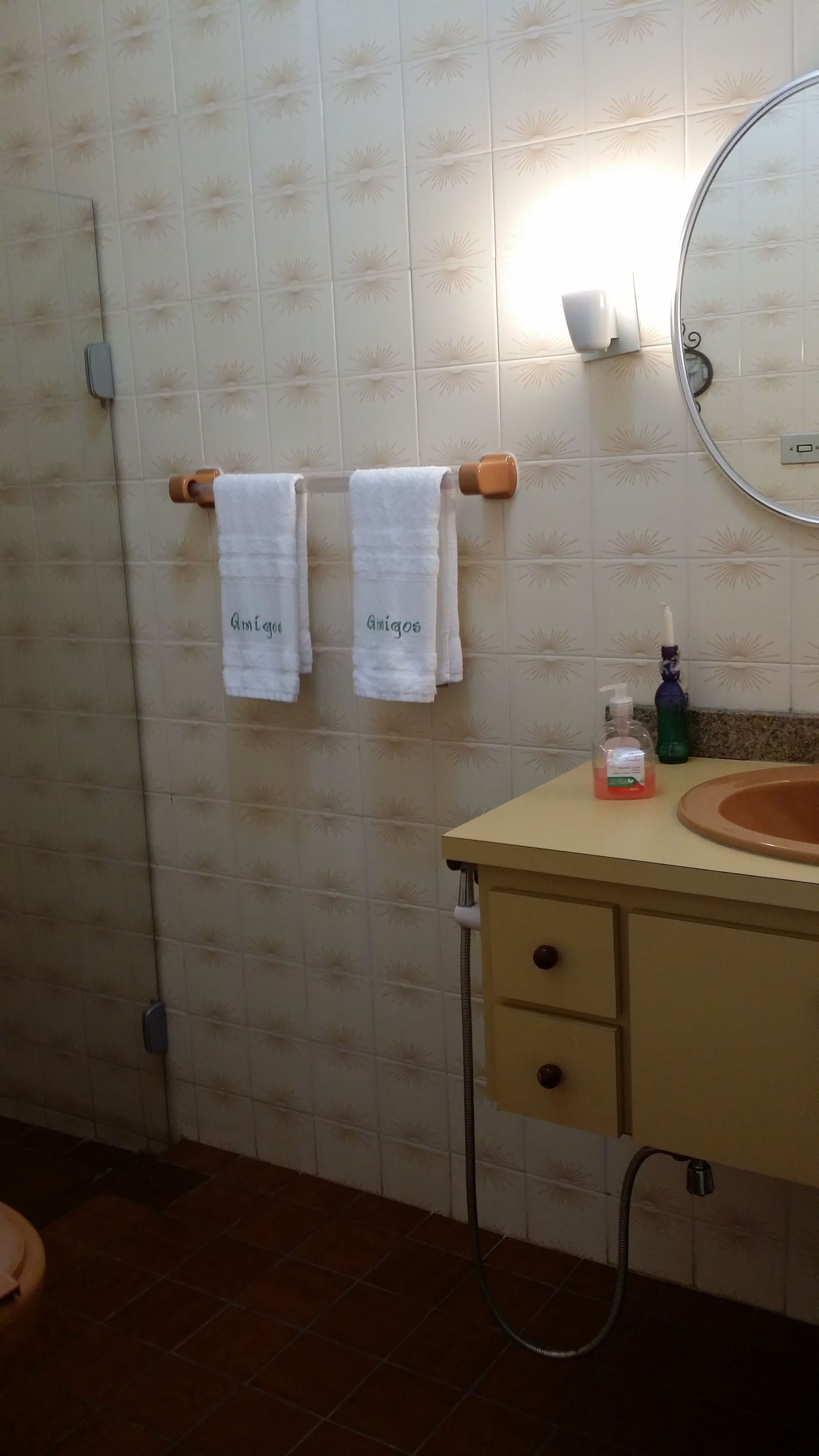 Un baño con pared de azulejos, dos toallas colgadas en un perchero, un lavamanos con espejo y un cabezal de ducha manual.