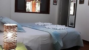 A bedroom with a bed, a bedside lamp, a television, and a window with blinds. - Suites em Águas de São Pedro (Aguas de Sao Pedro)