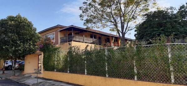 Una casa de dos pisos con balcón, rodeada de una cerca de alambre y follaje.