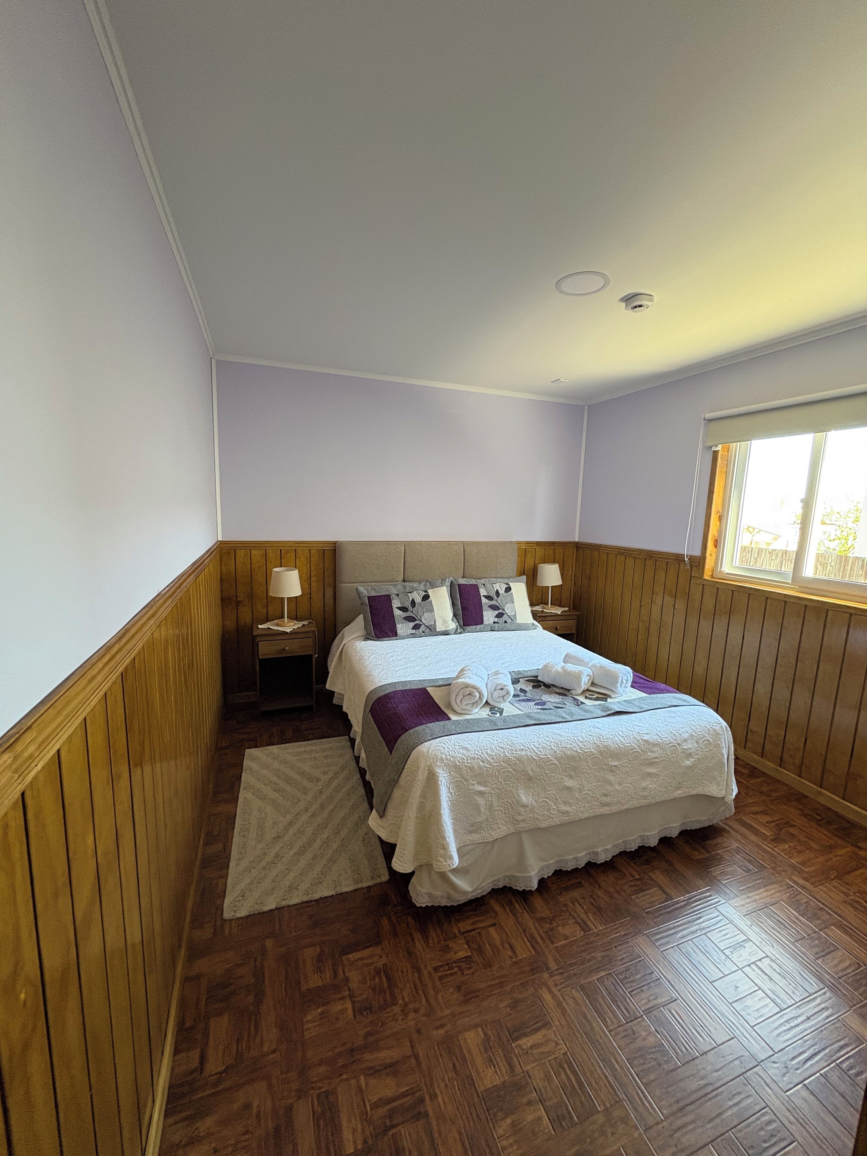 A bedroom with a wooden floor, a bed with white bedding and purple pillows, a nightstand with a lamp, and a window with blinds.