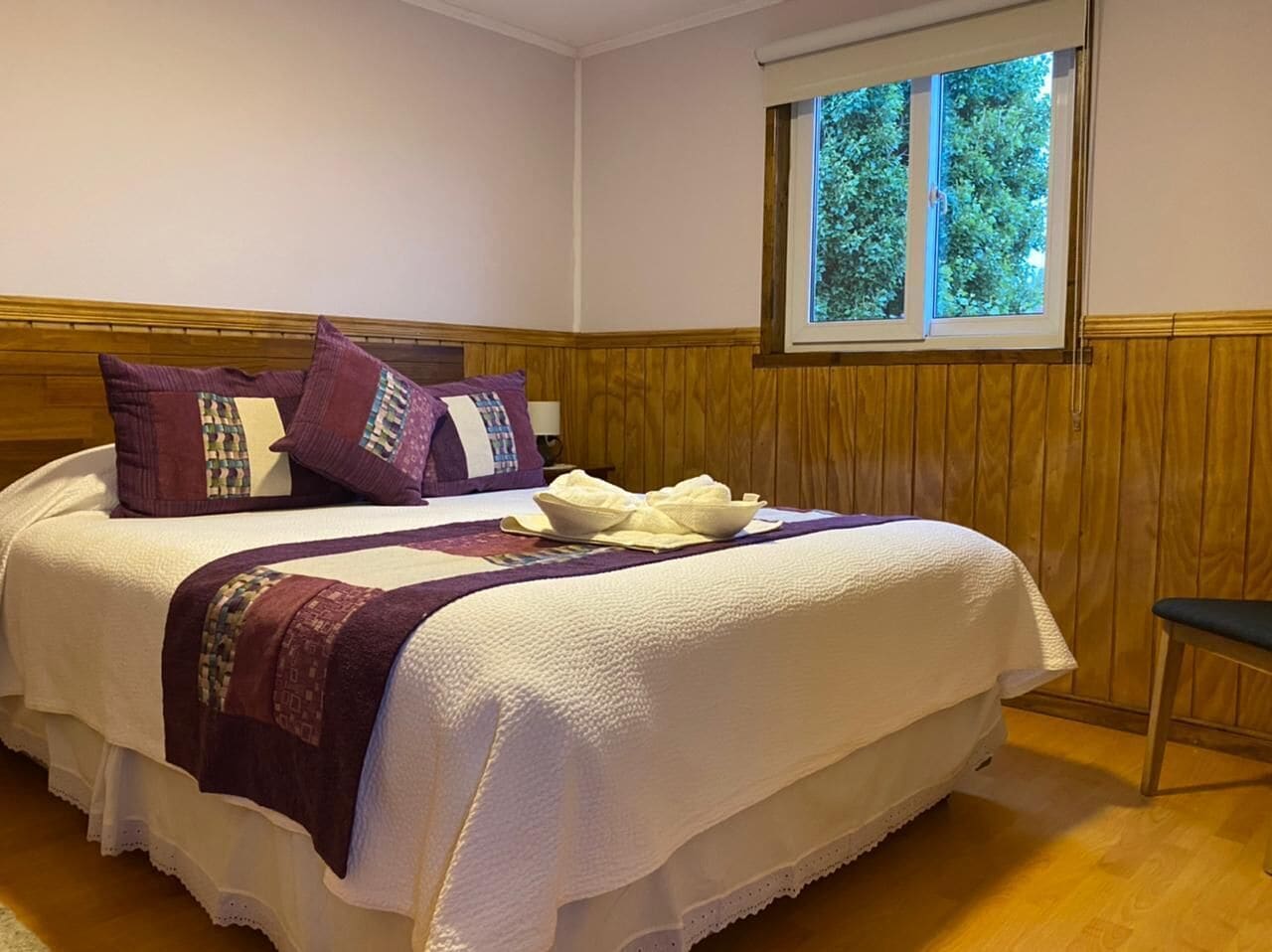Un lit bien fait, recouvert d’une couvre-lit blanche et violette, avec une tête de lit en bois et une fenêtre donnant sur des arbres.