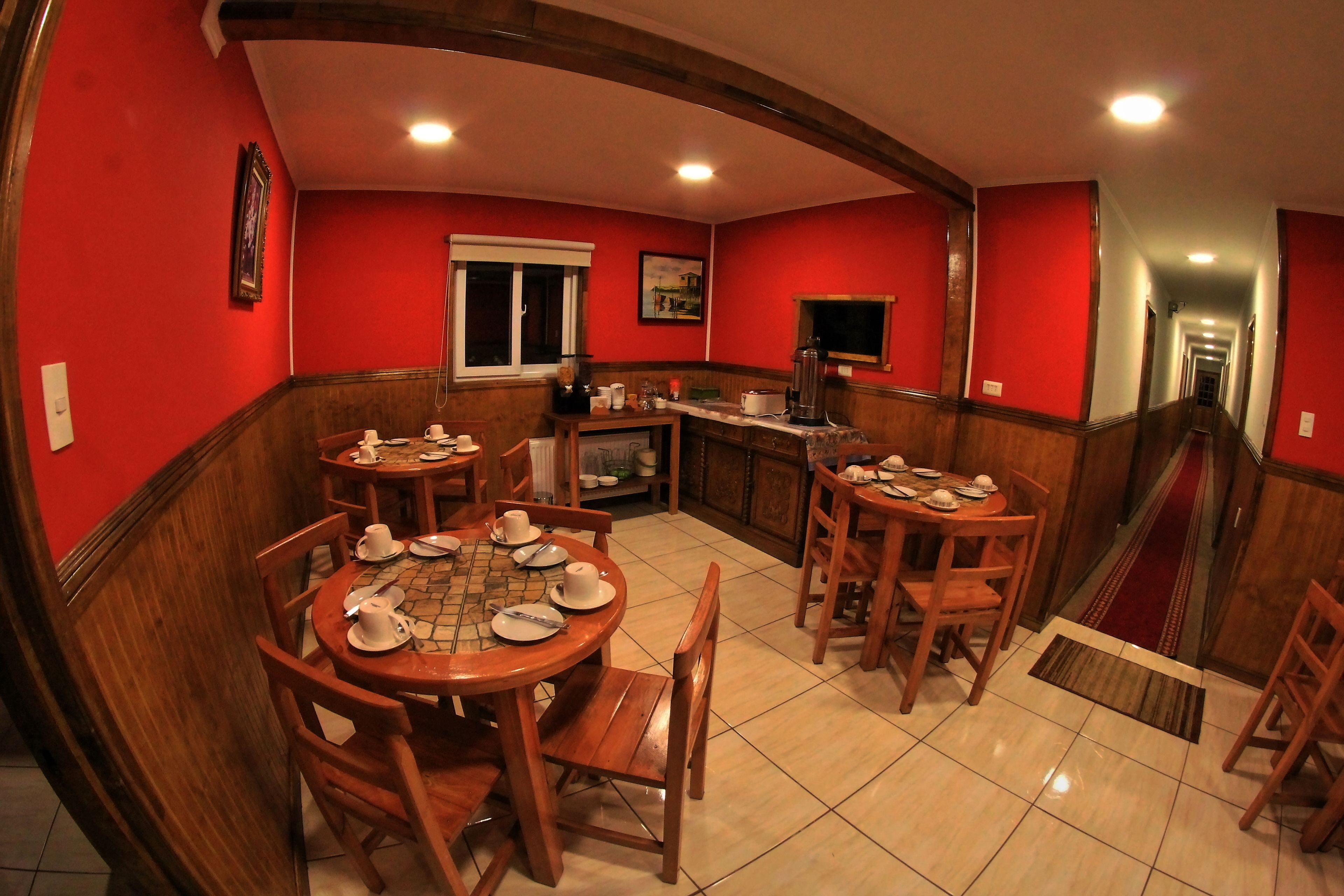Un comedor con mesas y sillas de madera, una cocina al fondo y una pared roja.