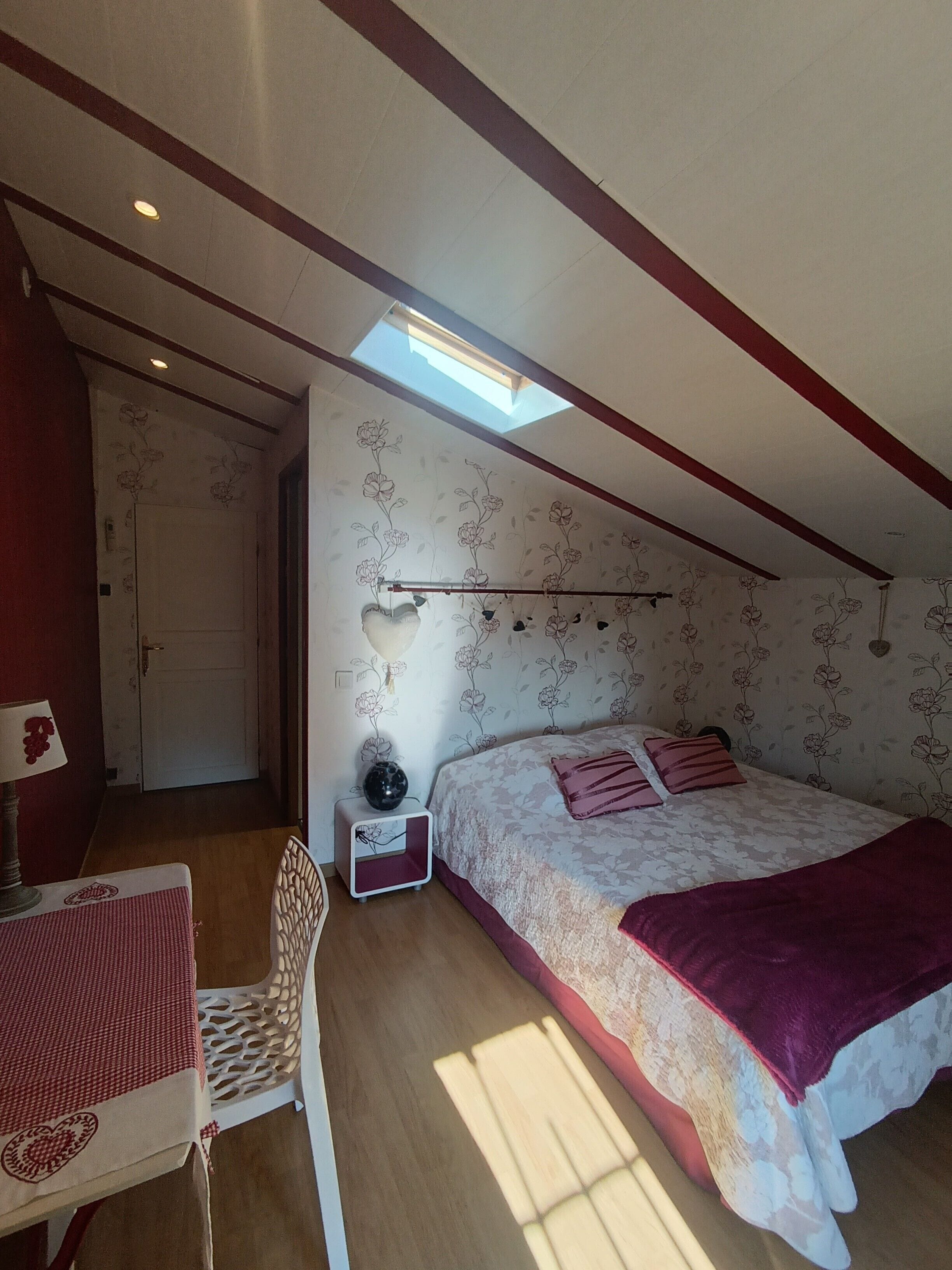 Un dormitorio con cama, una silla, una mesita, una mesita de noche y un tragaluz.