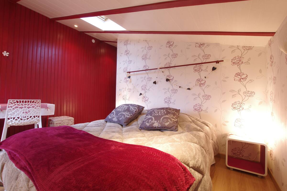 Een slaapkamer met een bed, kussens, een rode deken, een klein tafeltje met een kantkleed en een nachtkastje met bloemendecoratie.