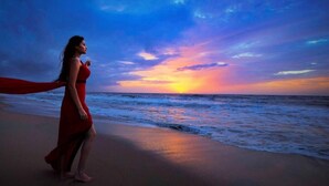 Une femme vêtue d’une robe rouge se promène sur une plage, avec un coucher de soleil en arrière-plan.